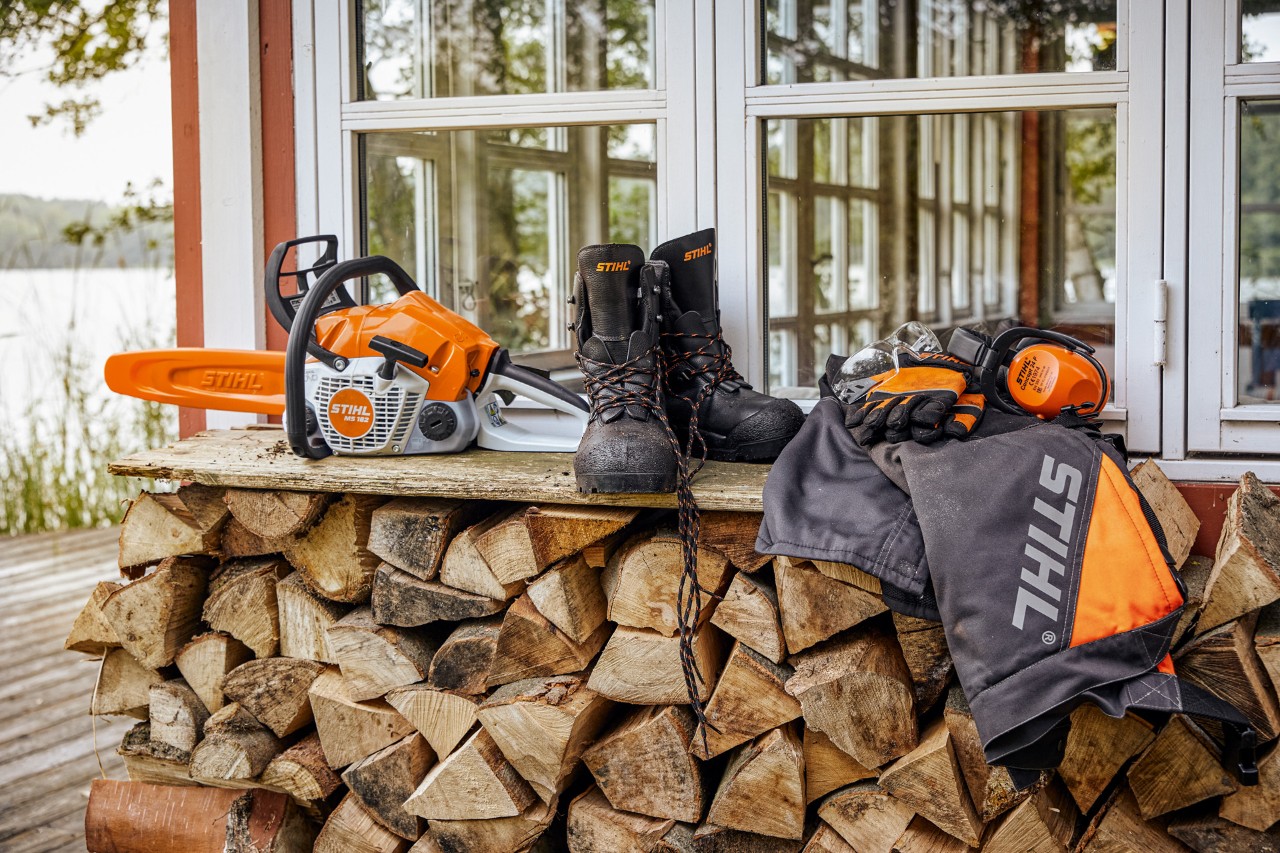 Odzież ochronna STIHL i MS 162 leżą na regale nad stosem drewna opałowego