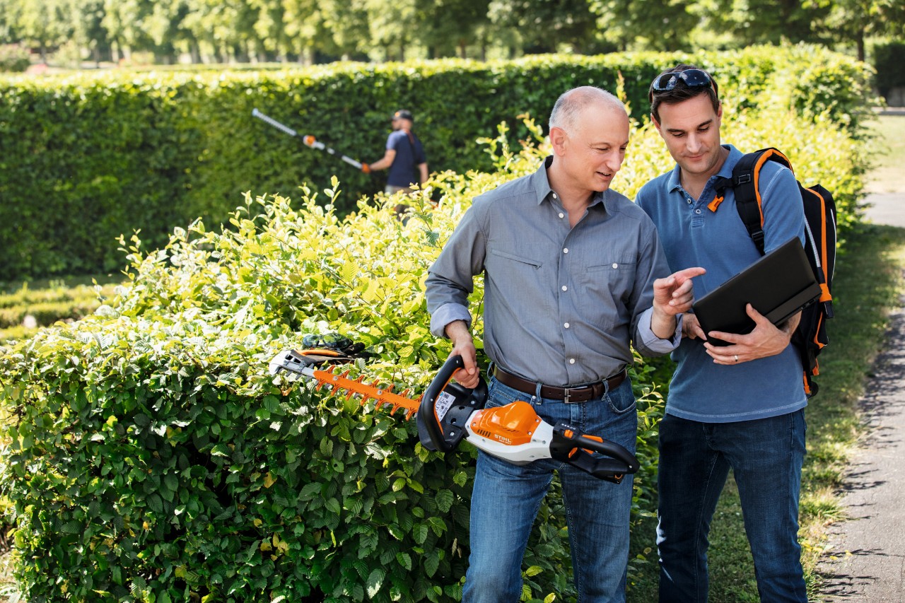 Dr Nikolas STIHL trzyma nożyce do żywopłotu STIHL i rozmawia z osobą stojącą obok trzymającą laptopa.