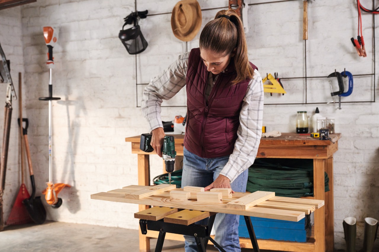 Kobieta w koszuli w kratkę i uczesana w kucyk przykręca listwy do poprzeczek ułożonych na stole roboczym, w tle ściana z urządzeniami