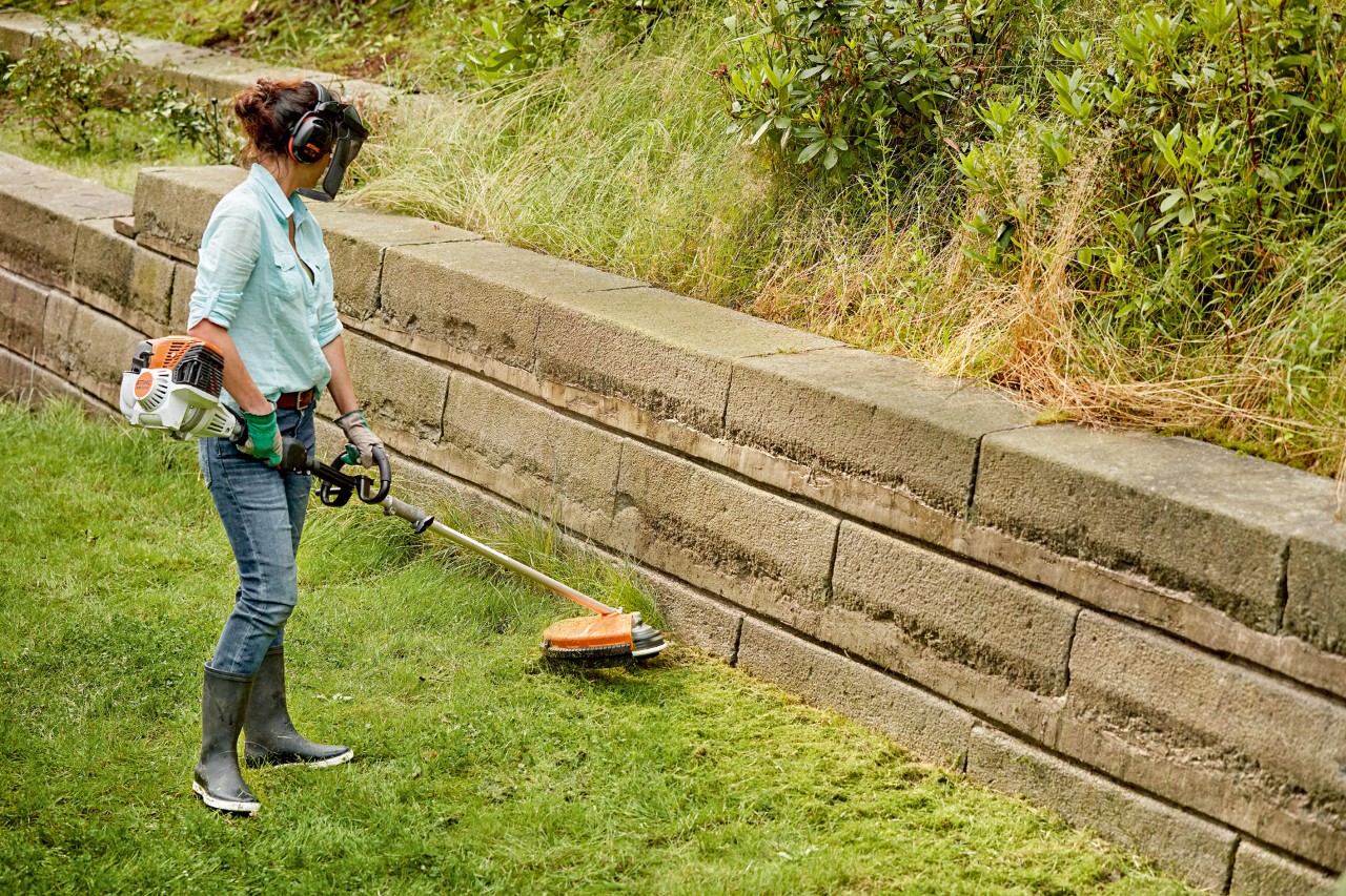 Kobieta w ochronie słuchu/twarzy tnie krawędzie trawnika przy ścianie kosą mechaniczną STIHL FS