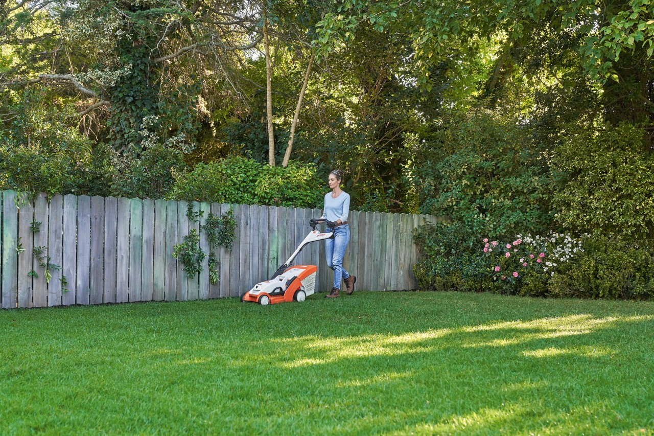 Kobieta kosi zielony trawnik przed drewnianym ogrodzeniem za pomocą kosiarki akumulatorowej STIHL RMA 339 C.