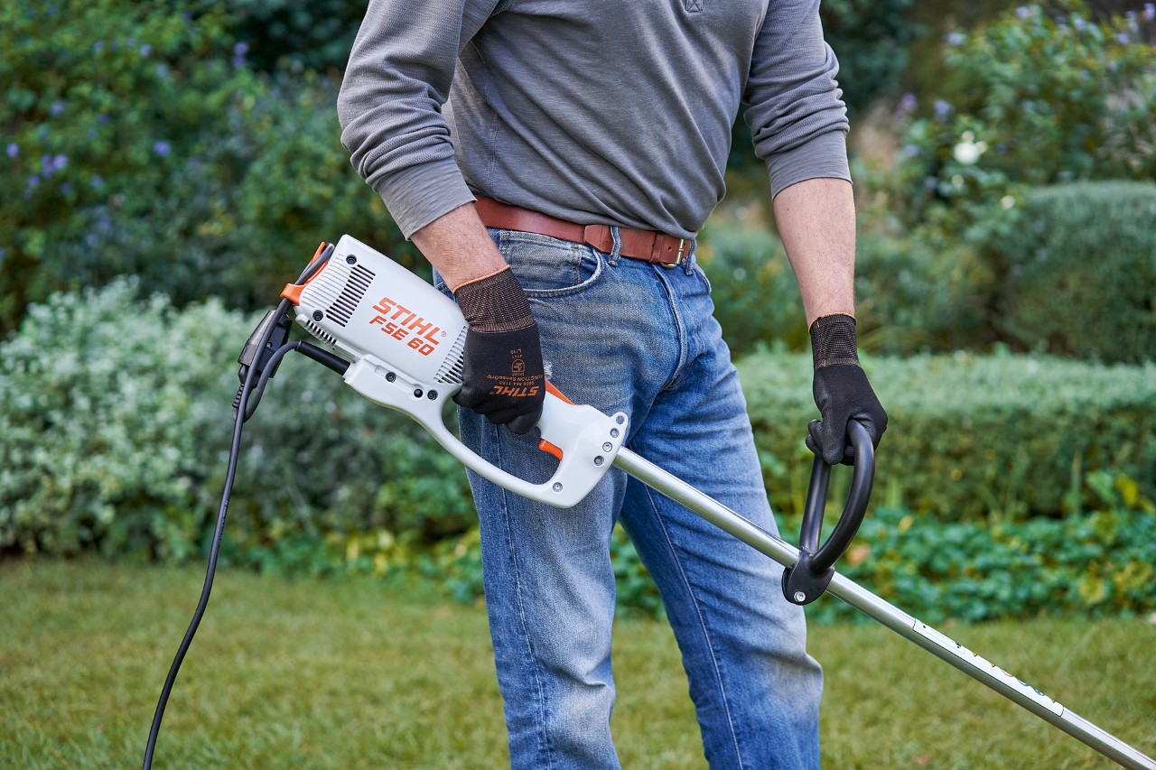 Mężczyzna w rękawicach ochronnych chwyta elektryczną kosę mechaniczną STIHL FSE 60 obiema rękami