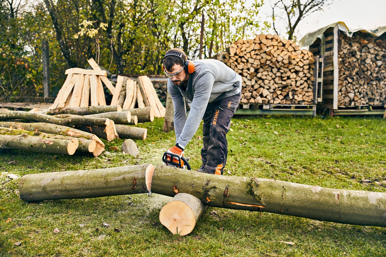 Mężczyzna w okularach ochronnych i ochronnikach słuchu przecina drewnianą kłodę spalinową pilarką łańcuchową STIHL MS 180
