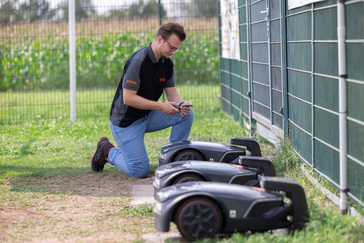 Kierownik ds. produktów STIHL iMOW® ,Thilo Ehrentreich, klęczy obok trzech robotów koszących z telefonem komórkowym w dłoni