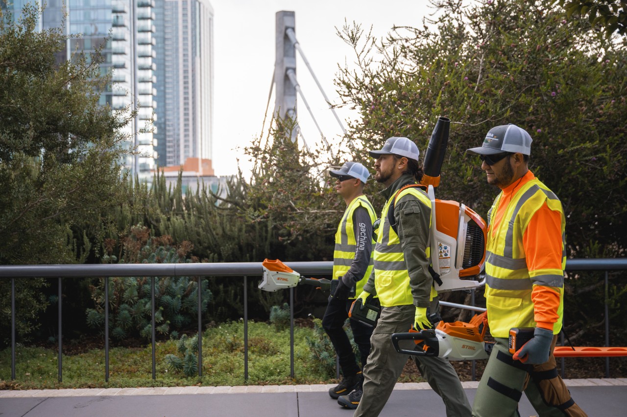 Trzech pracowników LaGa w odzieży roboczej i z urządzeniami STIHL przechodzi przez miasto.