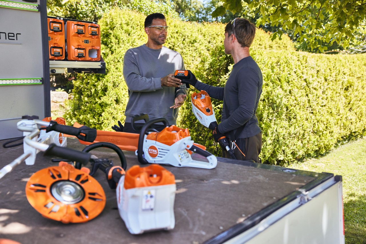 Mężczyzna przekazuje swojemu koledze akumulator STIHL, obaj znajdują się za powierzchnią ładunkową pojazdu, na której leży między innymi akumulatorowa pilarka spalinowa, bottTainer oraz inne urządzenia