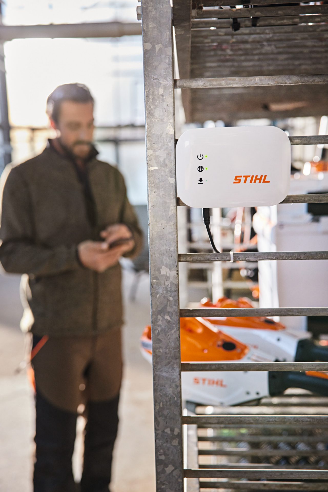 Skrzynka STIHL connected zawieszona na półce warsztatowej. W tle widać ogrodnika i architekta krajobrazu, który pracuje ze swoim smartfonem. 